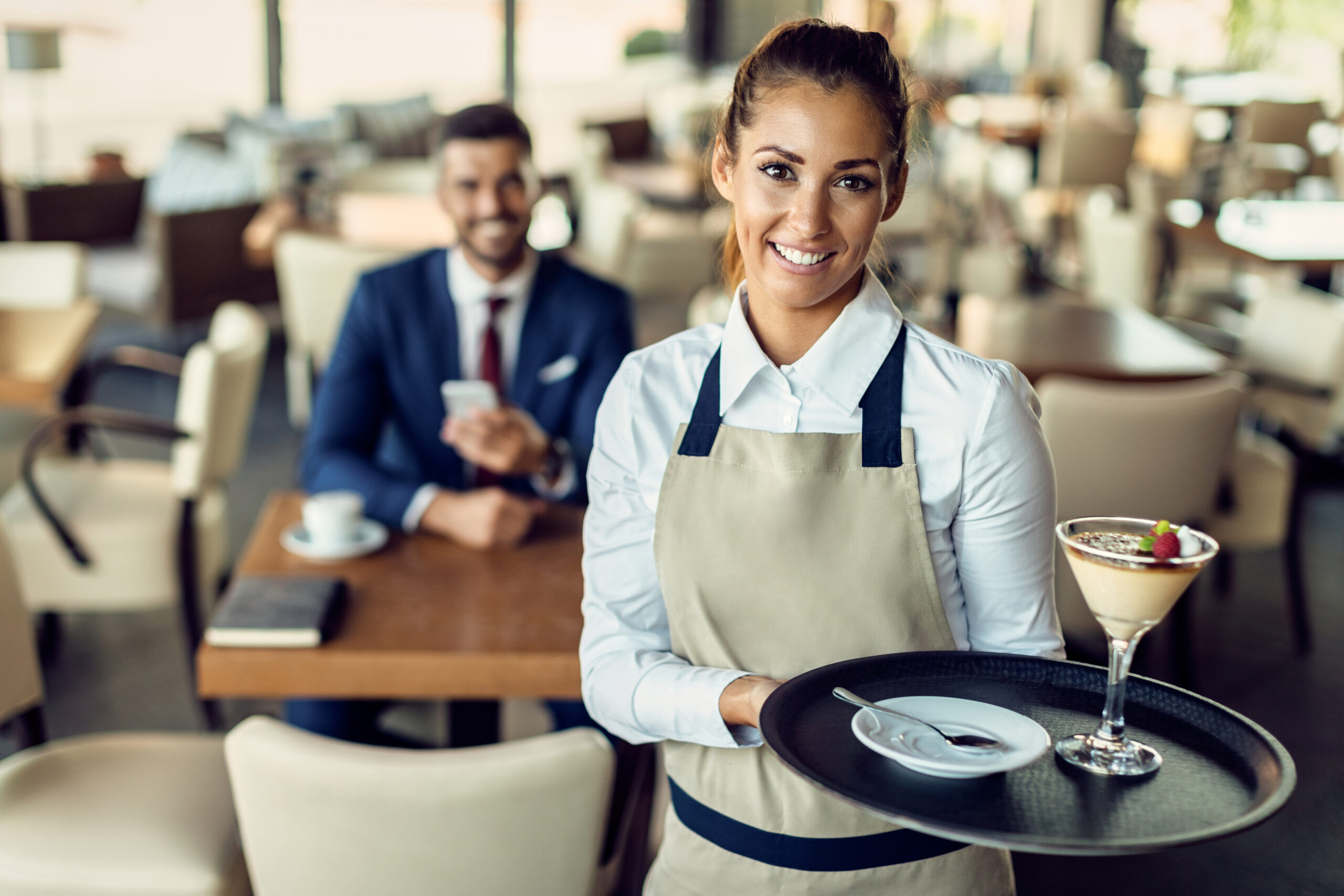 Portrait of happy waitress holding a tray with a lemon tart with raspberry sorbe and looking at camera. There is a businessman sitting at the table in the background.
